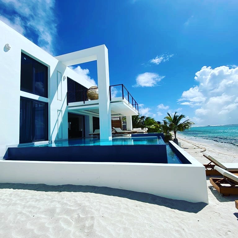 Luxury Bonaire villa with a modern white exterior, a private infinity pool on a white sand beach, and direct access to the turquoise Caribbean ocean under a blue sky.